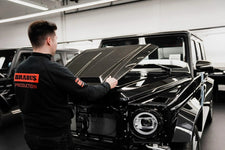 Person wearing a Brabus Production jacket inspecting a black luxury vehicle in a garage.