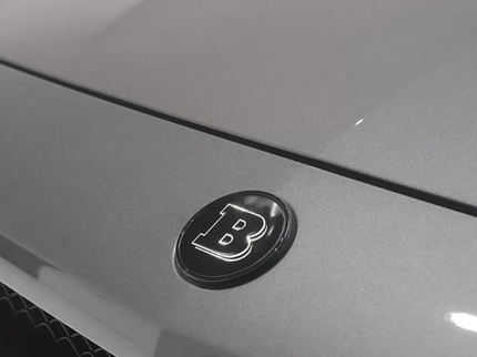 Close-up of a car emblem with a letter 'B' on a gray vehicle surface.